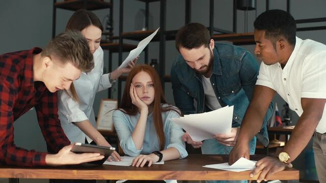 Portrait of exhausted young business woman sitting at table, looking at camera, multi-ethnic colleagues shaking paper documents. Multiethnic business team wants to get answer and decision from boss.