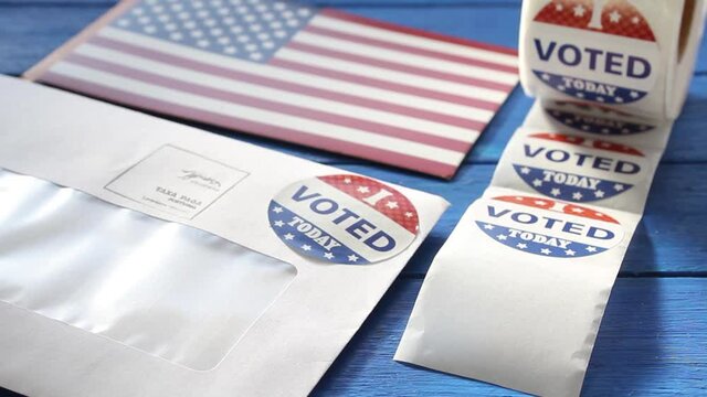 Woman's hand taking a I Voted today sticker from a roll on table with envelope and USA flag. Vote by mail in US presidential election concept
