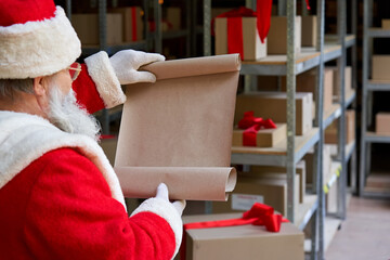 Over shoulder view of old Santa Claus wearing costume reading blank empty wish list standing in workshop gifts warehouse. Merry Christmas wishlist presents parcels post shipping delivery concept.