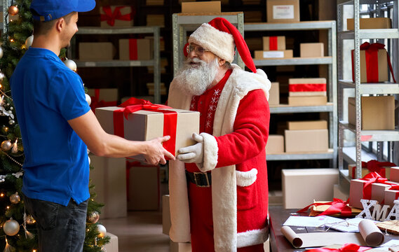 Happy old funny bearded Santa Claus wears costume holding Merry Christmas present giving gift box to courier worker delivering parcel standing in workshop. Xmas fast express shipping delivery concept