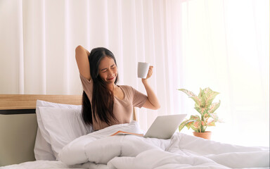 Lifestyle technology and working communication concept. Freelancer young woman sitting on the white bed with laptop checking social apps  while holding cup of coffee or tea in the morning at home.