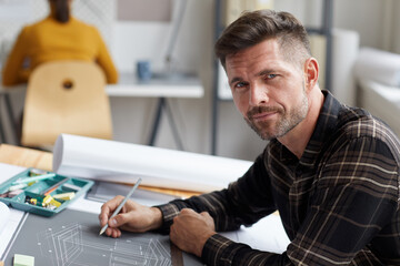 Portrait of mature bearded architect looking at camera while working on blueprints and plans sitting at drawing desk in office, copy space