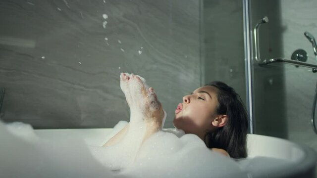 Smiling girl blowing foam in tub in bathroom. Beautiful woman taking bubble bath