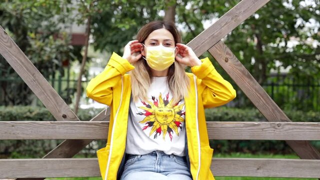 Young girl with yellow raincoat sitting at bench in park, wearing yellow medical mask and smiling to camera. Outdoor pandemic prevention in style. Attractive woman putting her face mask on. 