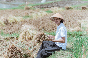 Naklejka na ściany i meble Male Asian farmer smiles wearing a hat looking at the camera while using a digital tablet after harvesting in the fields