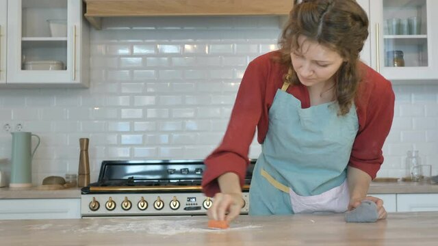 Unhappy young housewife in blue apron wipes wooden table covered with white flour using sponge in contemporary light kitchen
