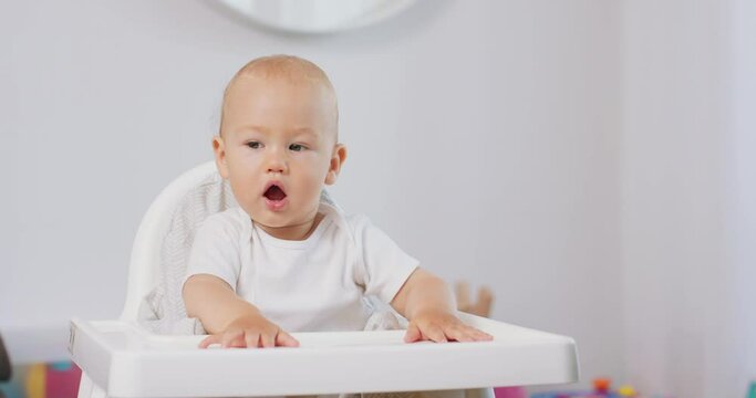 Portrait of young baby in white high baby chair. White walls on background.