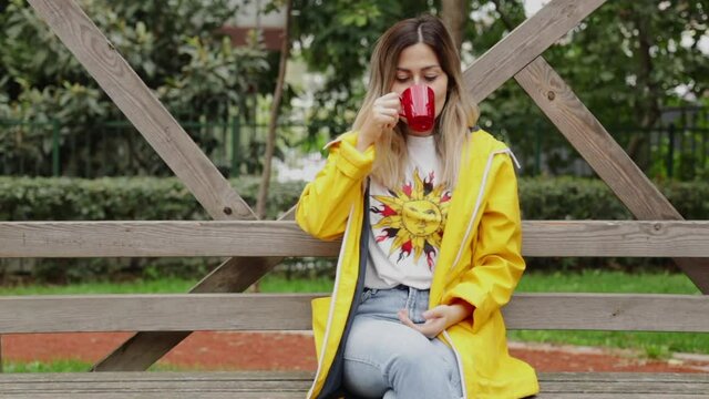 Beautiful young girl wearing yellow raincoat on a cloudy spring day sitting at park on bench, drinking coffee and looking to the camera, smiling. Slow motion video shot. 