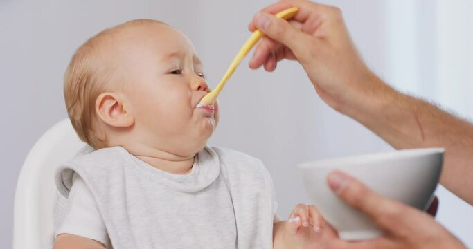 Closeup of young baby in white high baby chair and father's hand with a spoon and bowl, while he is feeding the baby, who eats and makes funny grimaces. White walls on background.