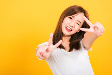 Asian happy portrait beautiful cute young woman teen smile standing wear t-shirt showing finger making v-sign symbol near eye  looking to camera isolated, studio shot yellow background with copy space