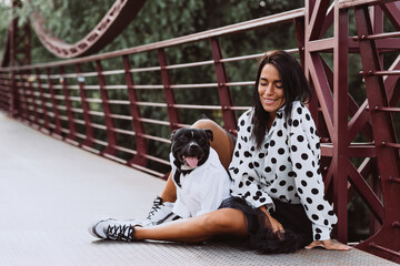 A beautiful young woman in a fluffy skirt and a polka-dot blouse sits on the bridge with her Staffordshire Bull Terrier dog. Soft selective focus. Artistic noise.