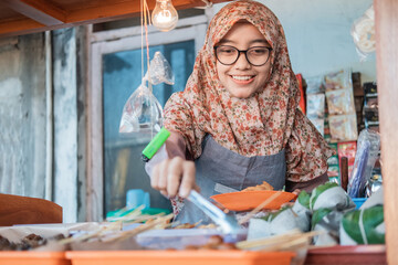 woman wearing a hijab, a stall seller, smiles while holding a food clip to tidy up the food displays in the cart stall