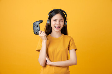 Cheerful beautiful Asian woman in casual yellow t-shirt and playing video games using joysticks with headphones on orange background. © sitthiphong