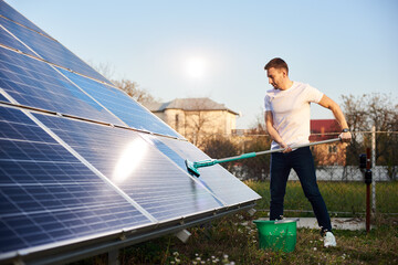 Young man is cleaning a solar panel with a mop, pv plant in rural area, side view