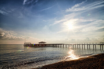 View from sand beach to water of sea, waves and pier in a nice day or evening with blues sky, bright sun and white clouds. The concept of a holiday on the sea or ocean in the South.