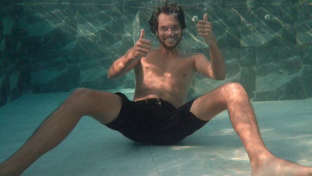 Underwater footage of joyful man on vacation diving into big swimming pool with thumbs-up