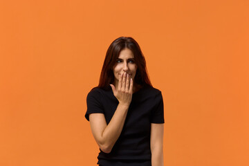 This is a dirty joke. Positive caucasian woman in black t-shirt covering mouth and feels shy hears hilarious jokes and giggles positively. Isolated over an orange wall