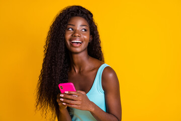 Photo portrait of young curly african american woman holding phone in two hands laughing wearing blue singlet isolated on bright yellow colored background