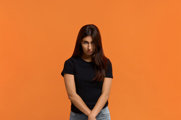 Obraz premium Forgive me. Miserable sad pleading caucasian woman in black t-shirt says sincere please, makes apology, keeps arms together in front of, begs with upset expression. Isolated over an orange wall
