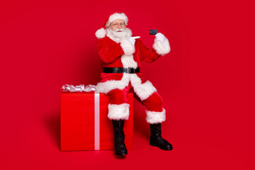 Full length body size view of his he handsome fat overweight cheerful Santa sitting on large big giftbox demonstrating plastic card isolated bright vivid shine vibrant red color background