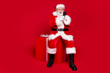 Full length body size view of his he nice handsome bearded cheerful Santa father sitting on big large giftbox chatting web shop order delivery isolated bright vivid shine vibrant red color background