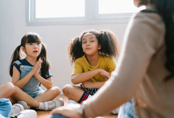 Elementary international students sitting on floor in circle around the teacher and listening a...