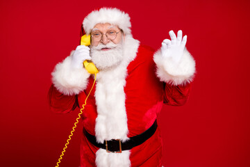 Photo of retired old man grey beard hold telephone show okey speak client solve conflict success wear x-mas costume leather belt gloves coat spectacles headwear isolated red color background