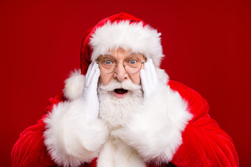 Photo of retired old man grey beard open mouth excited look hand cheekbone magical event deer start flying wear santa costume coat spectacles headwear isolated red color background