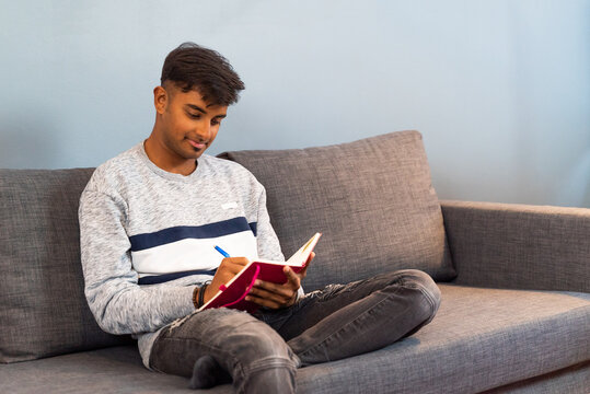Young Indian man happily writing at red book sitting at couch.