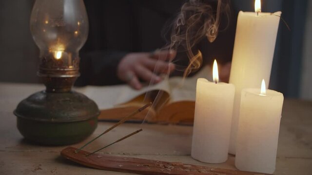Foreground burning candles and incense sticks placing on rustic table with blurry wizard in cape conjuring with vintage spellbook, casting rune stones while predicting future indoors in background.