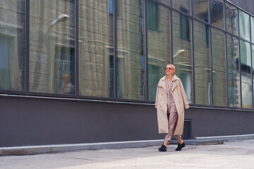 A woman with short hair in gray coat and glasses dancing, walking, smiling on a city street.