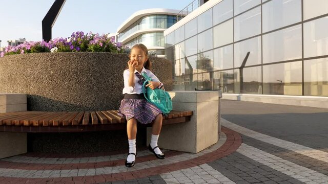 Little child girl with a backpack in school uniform is sitting on bench and eating the apple in school way.