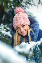 Portrait of beautiful smiling caucasian young woman standing in winter forest wearing blue jacket and pink hat