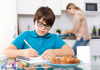 Naklejka na ściany i meble Positive schoolboy doing homework using laptop at kitchen interior, woman cooking at background