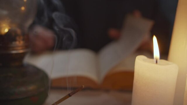 Close-up of burning candle lights and incense sticks standing on table with blurry monk in black robe running his finger under printed words of vintage book, studying ancient history in background.