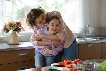 Loving Caucasian mom hug play with teen daughter cooking tasty salad for dinner at home kitchen....