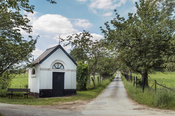 Obraz premium Chapel next to a road with trees. Bench next to it to rest and enjoy the view.