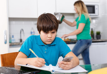 School age boy doing homework at kitchen while his mother preparing lunch at background