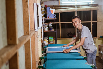 smiling asian boy brings and puts a silkscreen frame on top of the arranged t-shirt