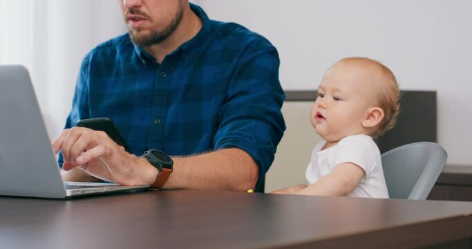 Baby at high baby chair, holds teether and pushes to the floor, hits the desk. Young bearded man sits at desk at home with laptop, writing on laptop keyboard, messaging on smartphone. Slow motion