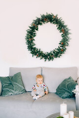 the child's first Christmas. a cute little boy is sitting in a festive costume on a gray sofa, against a white wall with a natural wreath of spruce.Christmas concept
