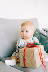 the child's first Christmas. a cute little boy is sitting in a festive costume on a gray sofa with a large gift with a red ribbon, against a white wall with a natural fir wreath.Christmas concept