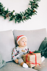 the child's first Christmas.brooding little boy sits in Santa hat and holiday costume on gray sofa with a large gift with red ribbon,against white wall with natural fir wreath.Christmas concept