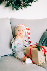 Obraz na płótnie Canvas the child's first Christmas.brooding little boy sits in Santa hat and holiday costume on gray sofa with a large gift with red ribbon,against white wall with natural fir wreath.Christmas concept