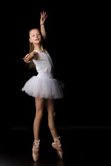 Naklejka premium Cute little girl in a tutu and pointe shoes is dancing in the studio on a black background.
