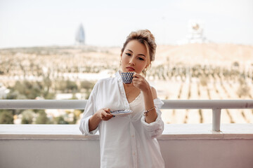 young beautiful Asian girl in beige lace shorts,white shirt drinking coffee standing on the balcony. selective focus. small focus area.