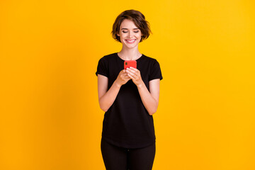 Portrait of attractive focused cheerful girl holding in hands using device chatting isolated over bright yellow color background