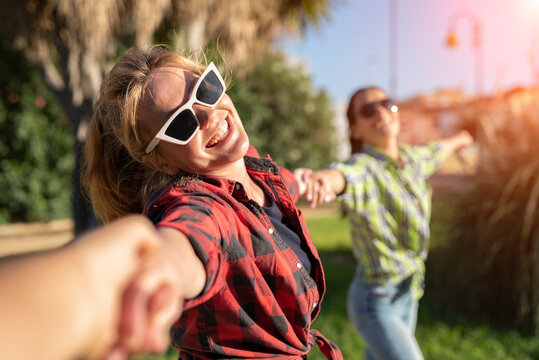 Pretty Young Caucasian Woman In Sunglasses Pulling Along By The Hand Invitingly With A Happy Smile As Her Friend On The Background. Sunny Summer Day