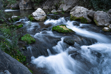 Fototapeta na wymiar Fast running mountain river with mossy boulders and rocks at cloudy day on long exposure.