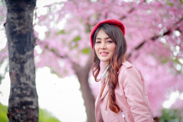 A traveler hipster woman sightseeing wear a red hat and a smooth leather dress with beautiful sakura cherry blossoms tree full blooming in pink color in the park on a spring day.